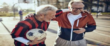 img title An older men laughing on a basketball court with a soccer ball in one hand and his arm around another laughing man