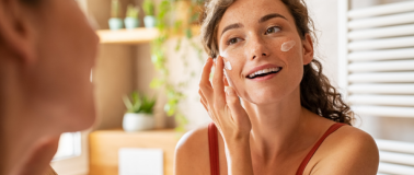 A woman uses her reflection in a mirror to apply facial moisturizer